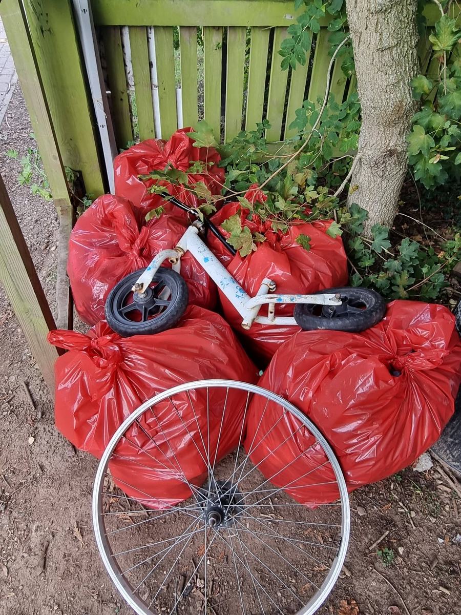 Red rubbish sacks stacked against a green fence, with a child's balance bike and a bicycle wheel in front