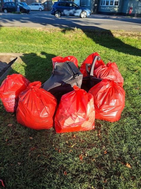A pile of red rubbish sacks and a broken black office chair on a grass verge beside a road