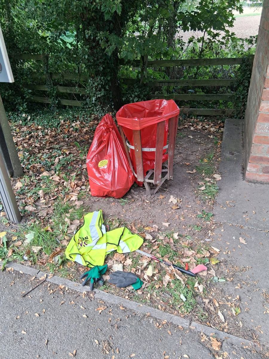 A hi-vis vest, a litter picker and two gloves on the ground beside a full red sack and a red bag in a wooden holder